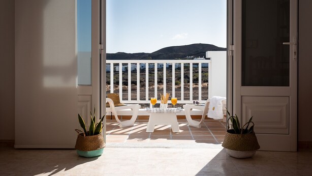 Un balcón con mesa y sillas, dos macetas con plantas y vista al mar y las colinas.