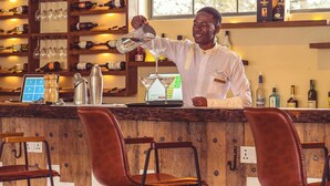 A bar area with a bartender pouring a drink, shelves with bottles, and a laptop on the counter. - Kili Seasons Hotel (Arusha)