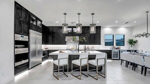 Une cuisine moderne avec des meubles de couleur sombre, des appareils en acier inoxydable, un îlot central doté de chaises, un éclairage suspendu et une vue sur la ville grâce à de grandes fenêtres.