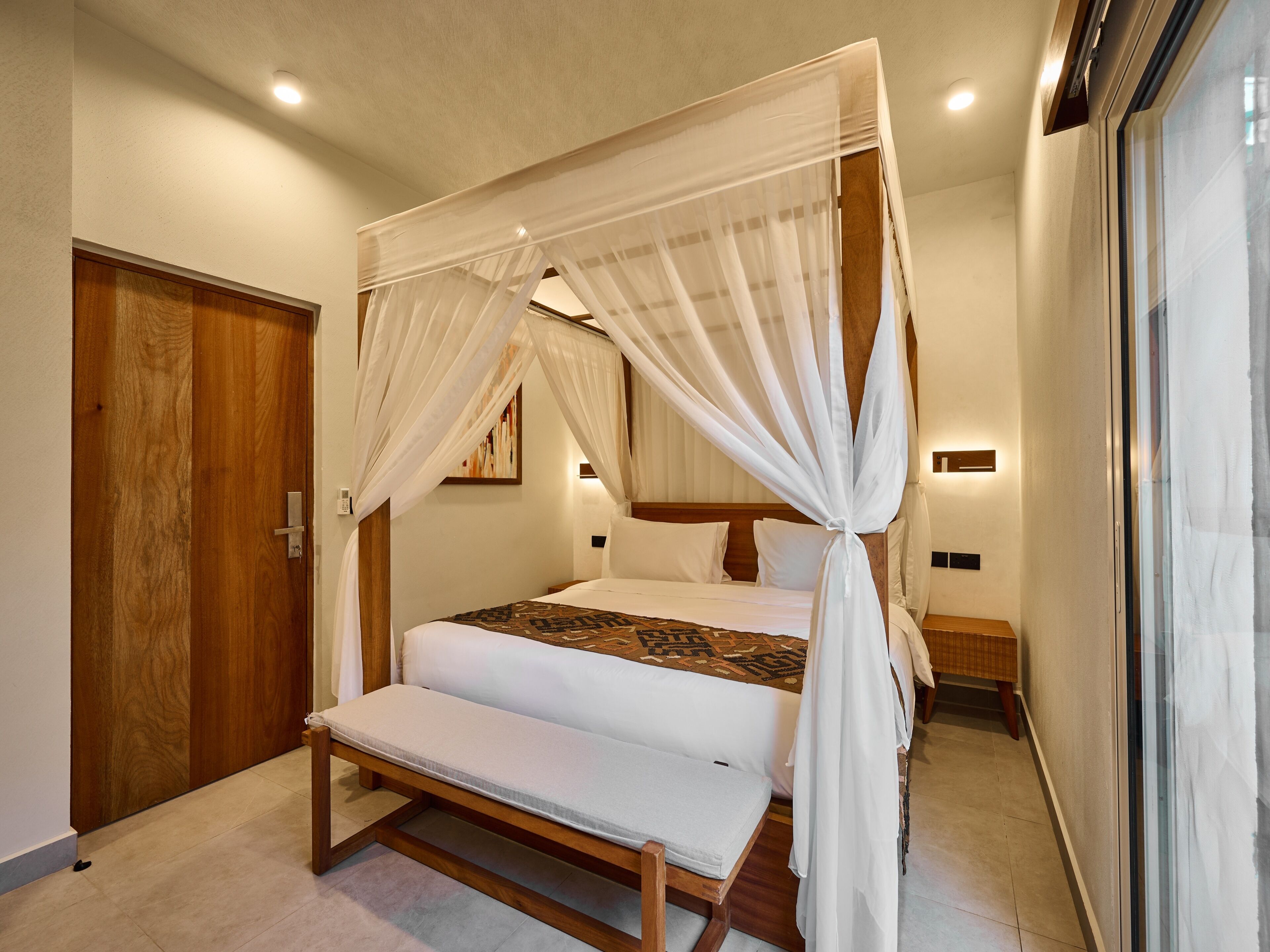 A bedroom with a four-poster bed, a bench, a wooden door, and a nightstand.