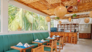 A restaurant with a green sofa and wooden tables, a bar area, and a view of palm trees. - Samawa Living (Paje)