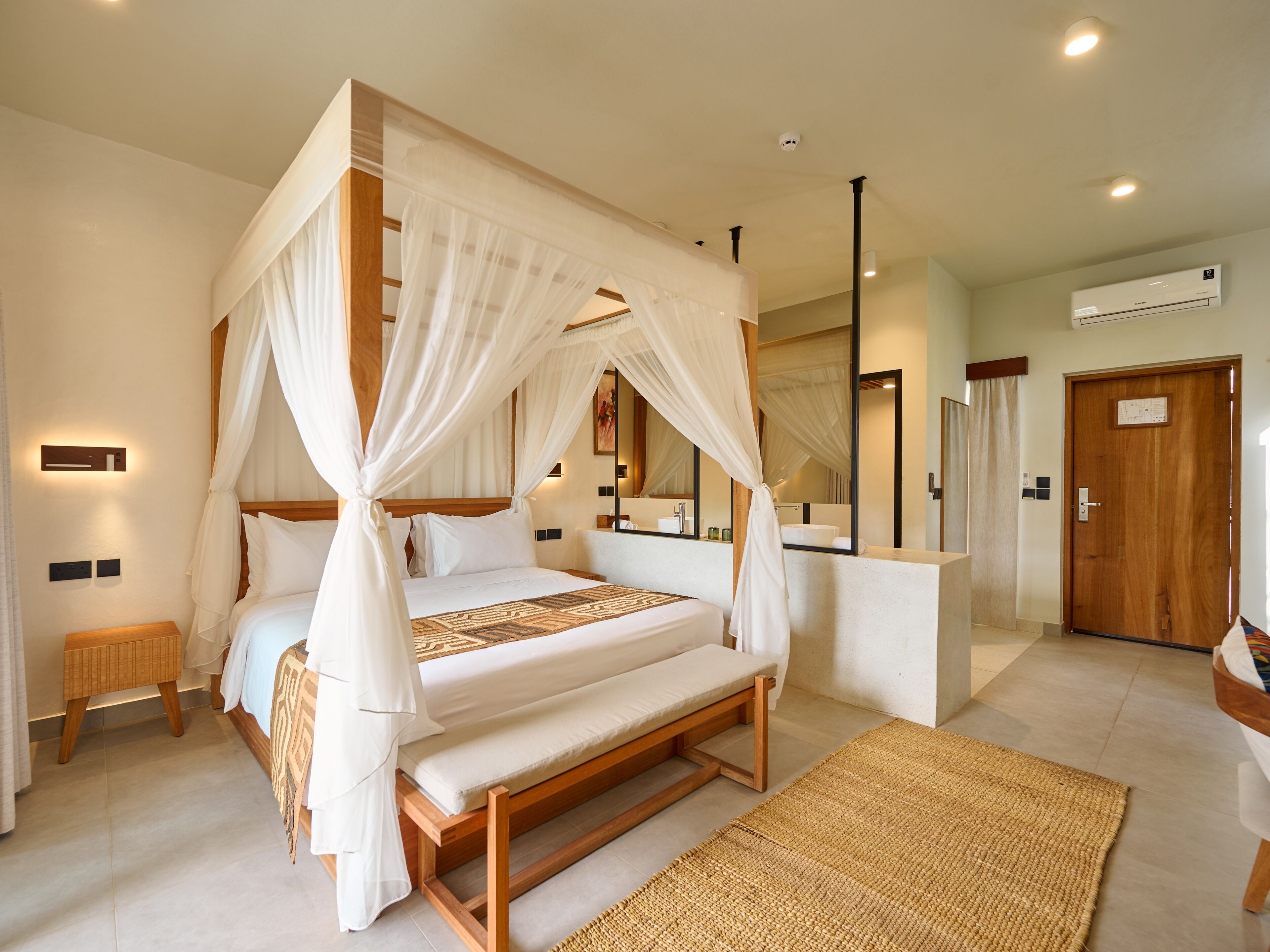 A four-poster bed with a canopy, a wooden bench, a woven rug, a bathroom with a mirror, and an air conditioning unit.
