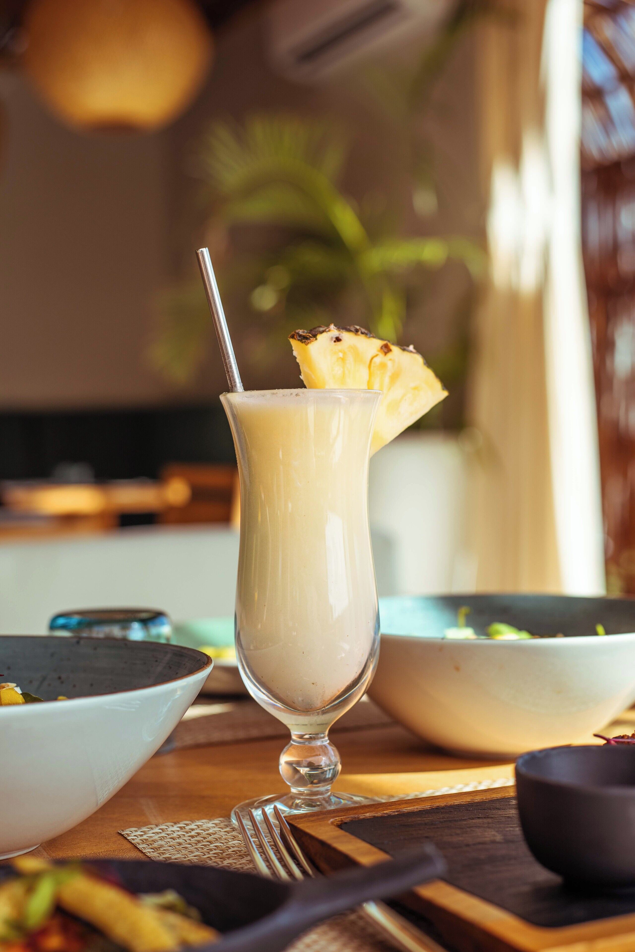 A glass of a light-colored beverage with a straw and a slice of pineapple on the rim, placed on a wooden table with a bowl and a spoon nearby.