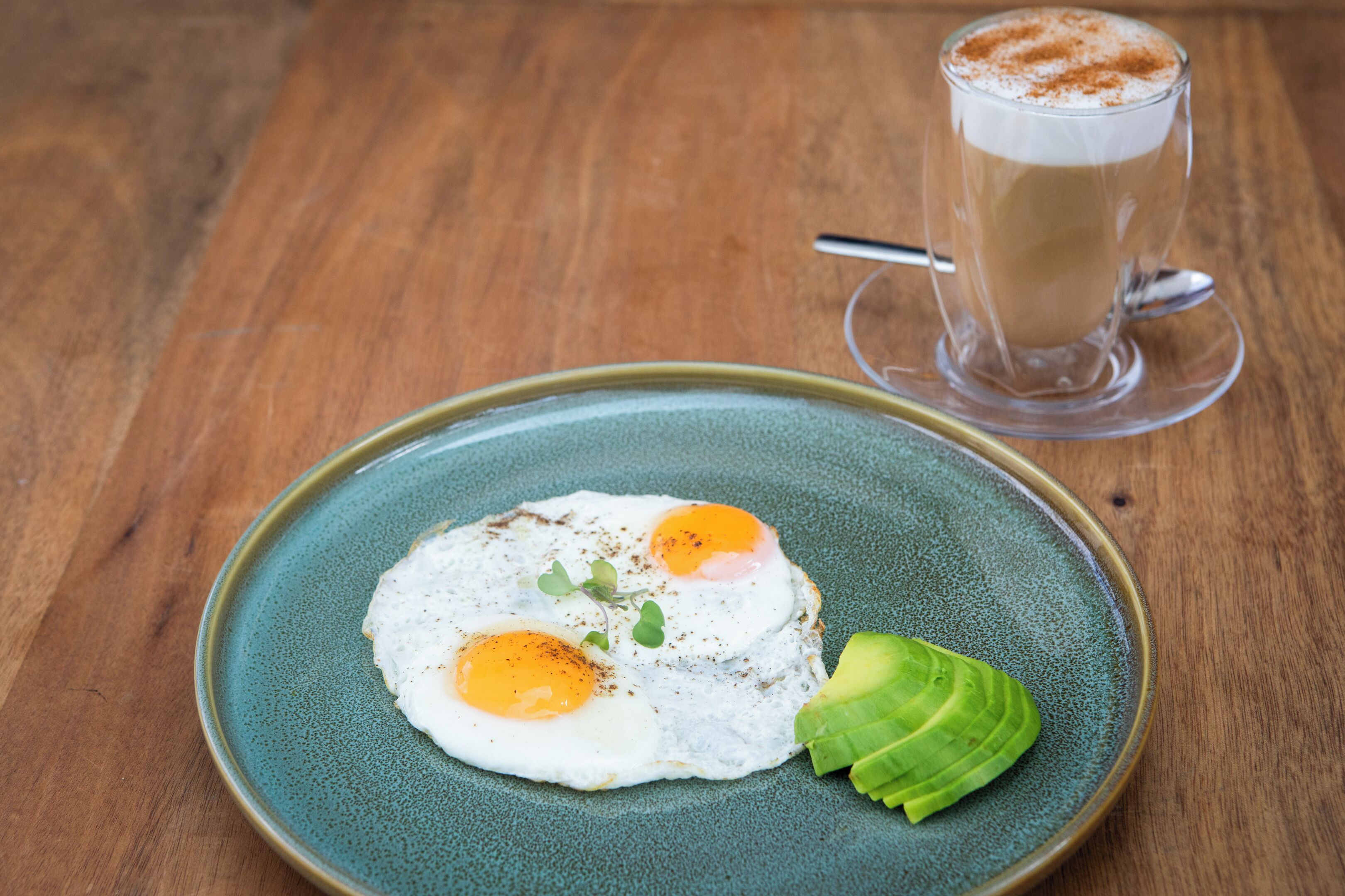 A plate with two sunny-side-up eggs and avocado on a ceramic plate, and a glass of coffee with a spoon on a wooden table.