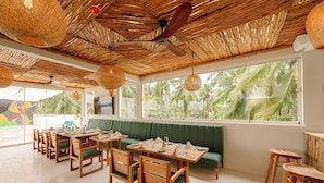 A restaurant with a thatched roof, wooden furniture, and large windows offering a view of palm trees. - Samawa Living (Paje)