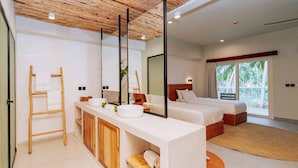 A modern bedroom with a large bed, a bedside table, a built-in wardrobe, and a bathroom with a sink and mirror. - Samawa Living (Paje)