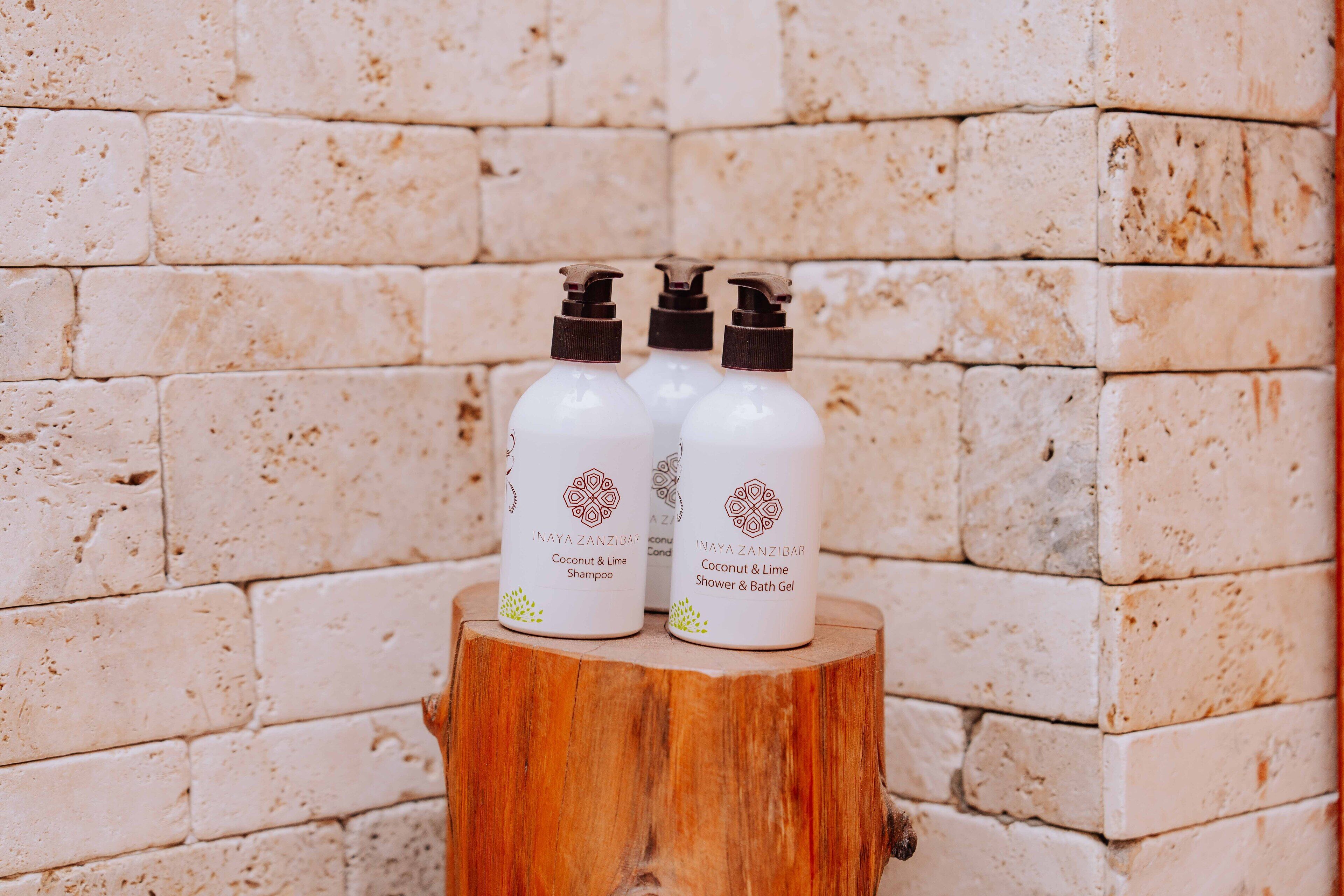 Drei Flaschen Inaya Zanzibar Kokos & Limette Shampoo und Dusch- und BadeGel auf einem Holzstumpf vor einer Ziegelwand.