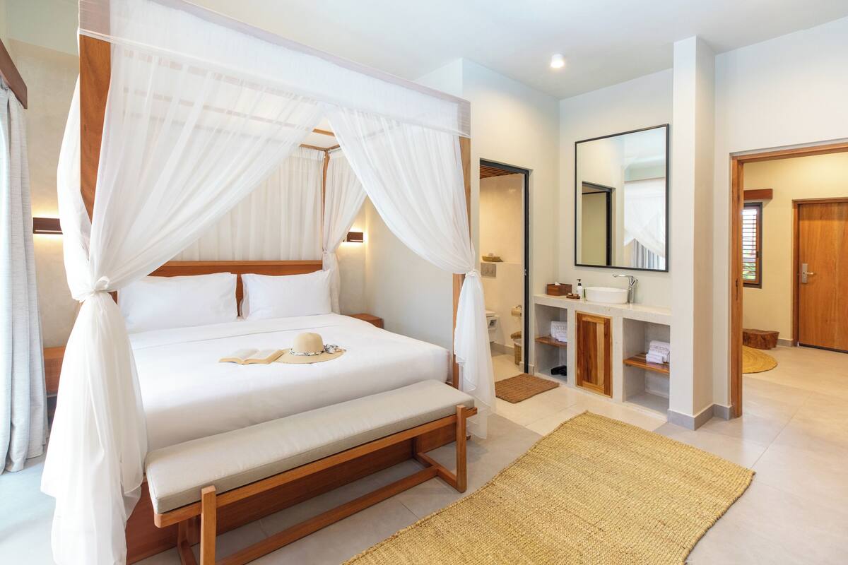 Ein geräumiges Schlafzimmer mit einem großen Bett, einem Nachttisch, einer Lampe, einer hölzernen Bank, einem Teppich und einem Badezimmer mit Waschbecken, Spiegel und Holztür.