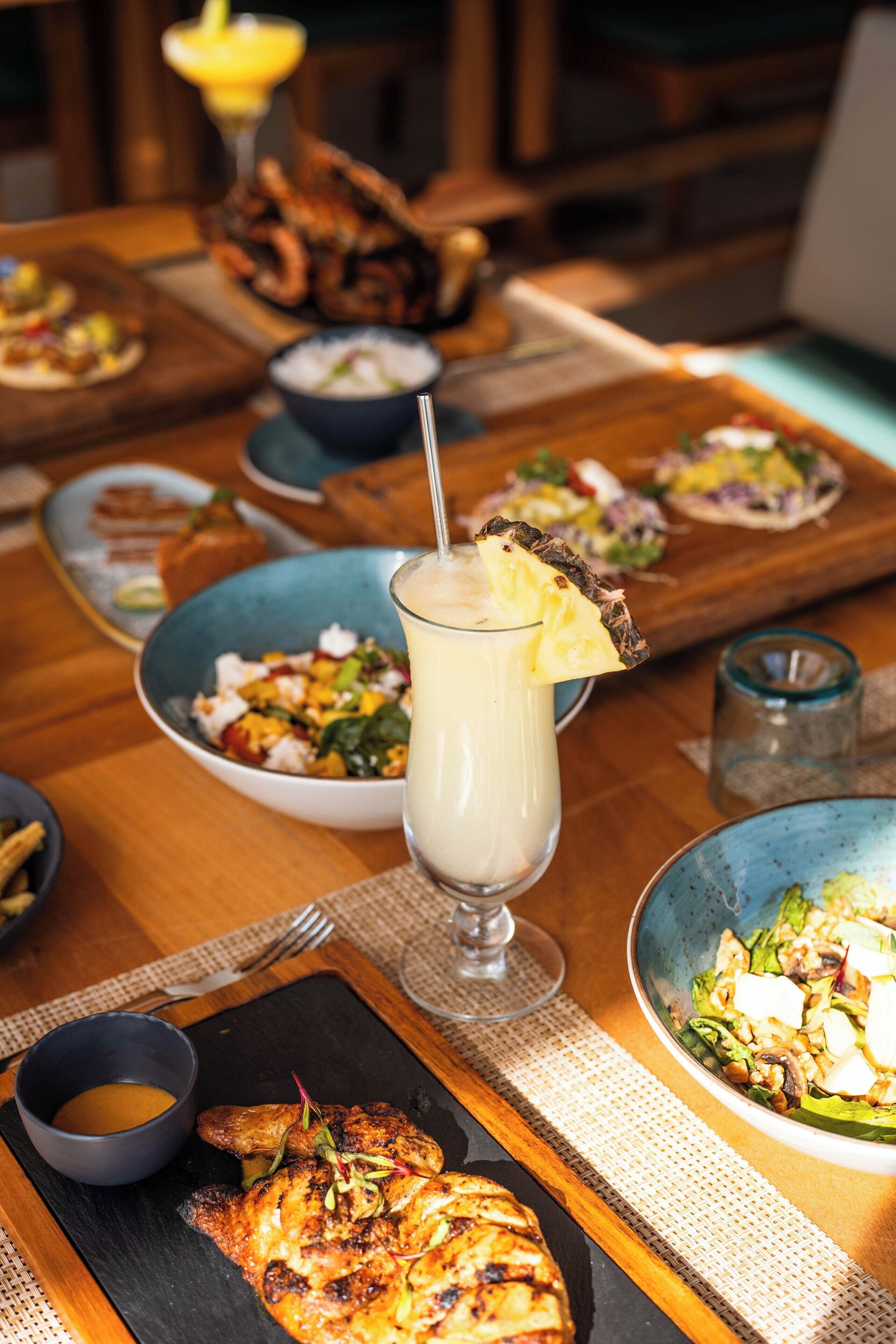 A dining table with various dishes, including grilled chicken, salads, and a milkshake with a pineapple garnish.