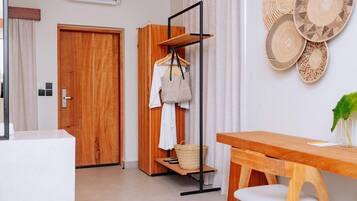 Una habitación con puerta de madera, un armario, una percha con ropa y una cesta, una mesa de madera con una silla y decoración mural tejida.