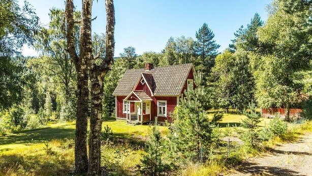 Une cabane en bois rouge, surmontée d’un toit en tuiles, entourée d’une forêt dense et sous un ciel dégagé.