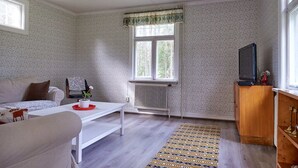 Un salon avec un canapé blanc, une table basse blanche, un tapis à motifs, un radiateur, une fenêtre agrémentée d’une lambrequin à motifs et un téléviseur posé sur un meuble en bois.