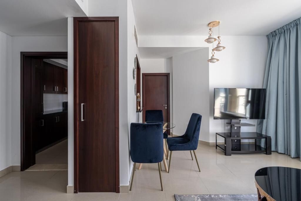A modern living room with a dark wooden door, a TV on a stand, two blue chairs, and a dining table.