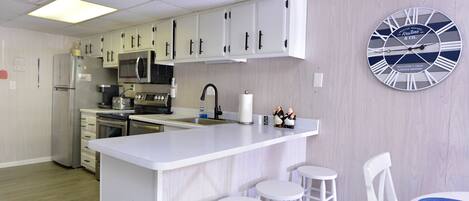 A modern kitchen with white cabinetry, a central island with stools, a large wall clock, and a microwave.