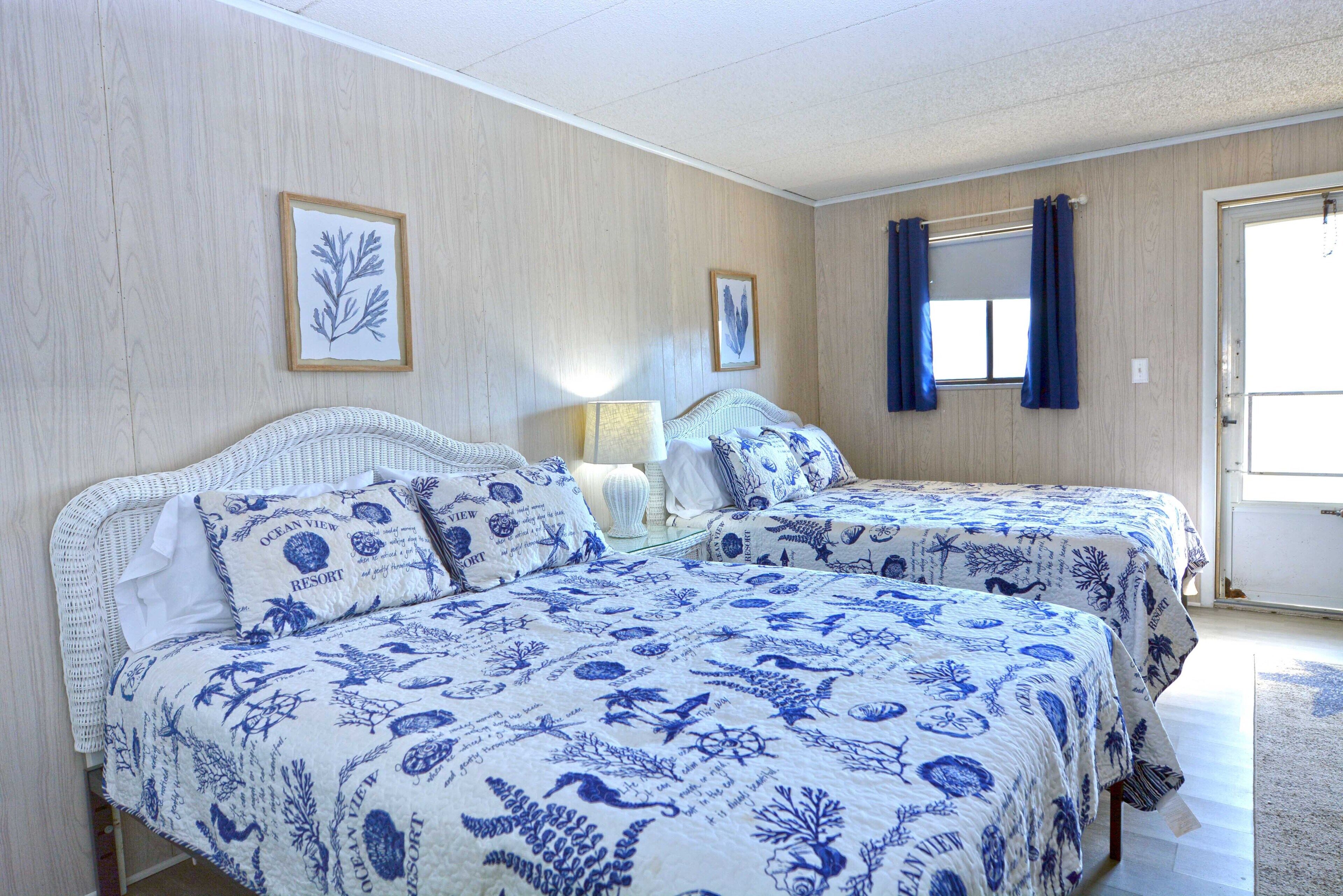 A bedroom with two beds, each with a blue and white ocean-themed bedspread, a white wicker headboard, and a framed picture on the wall.