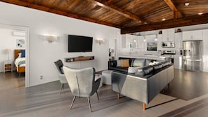 Un salon moderne avec un canapé gris, une table basse et un téléviseur fixé au mur. On y trouve également un coin cuisine avec des meubles blancs et des appareils électroménagers en acier inoxydable.