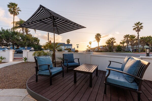 Une terrasse extérieure avec une plateforme en bois, deux chaises bleues rembourrées, une petite table et un parasol à rayures.
