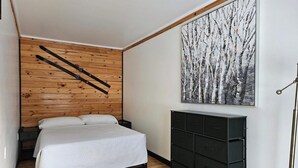 Makuuhuoneessa on puusäleistä tehty seinä, valkoisilla tekstiileillä varustettu sänky, musta lipasto ja suuri maalaus, jossa on kuvattuna puita.