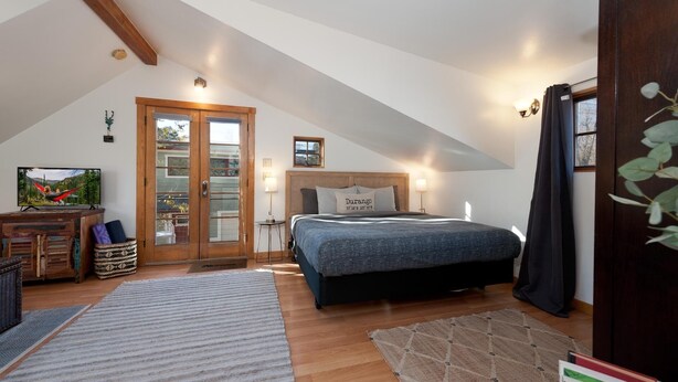 Ein Schlafzimmer mit schrägem Dach, einem Bett mit Kopfteil, einer Holztür mit Fensterfeldern, einem Fernseher und einem kleinen Tisch mit Korb.