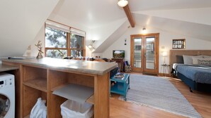 Ein gemütliches Schlafzimmer im Loft-Stil mit großem Fenster, einem Holztisch, einer Waschmaschine und einem Bett mit Kopfteil.