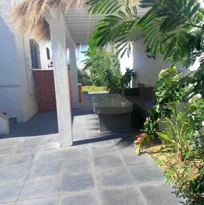Een overdekt buitengebied met een stenen vloer, een betonnen bank en diverse planten.