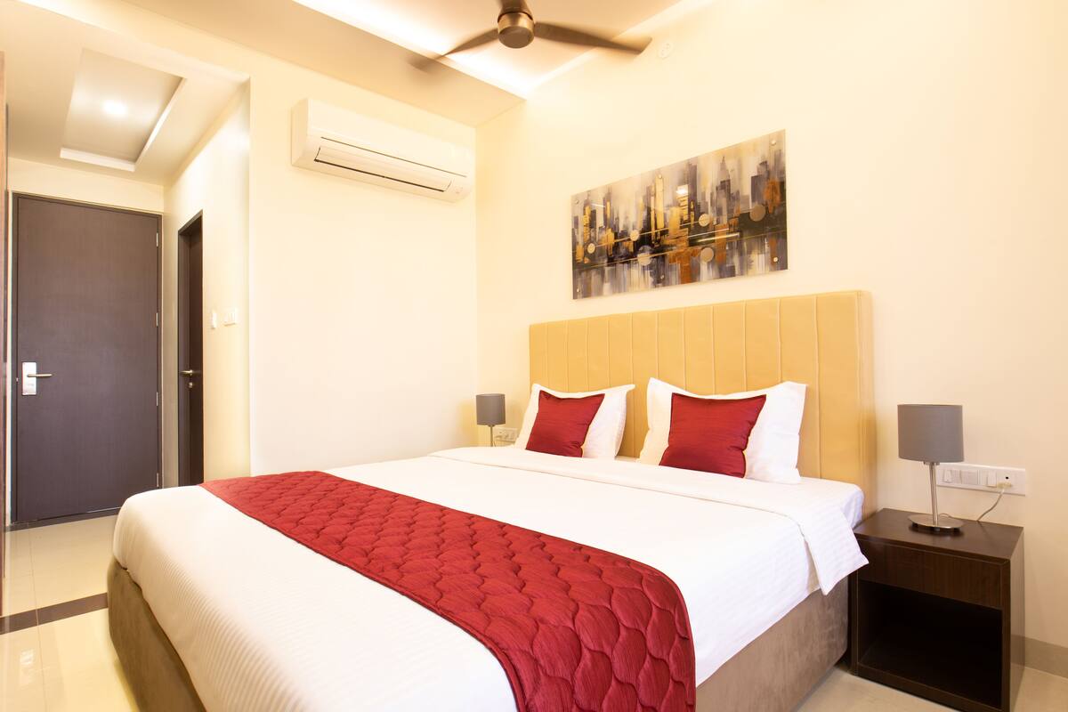 A hotel room with a bed, a nightstand, an air conditioner, a painting, and a door.