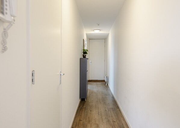 A narrow hallway with white walls and wooden flooring, leading to a door at the end.