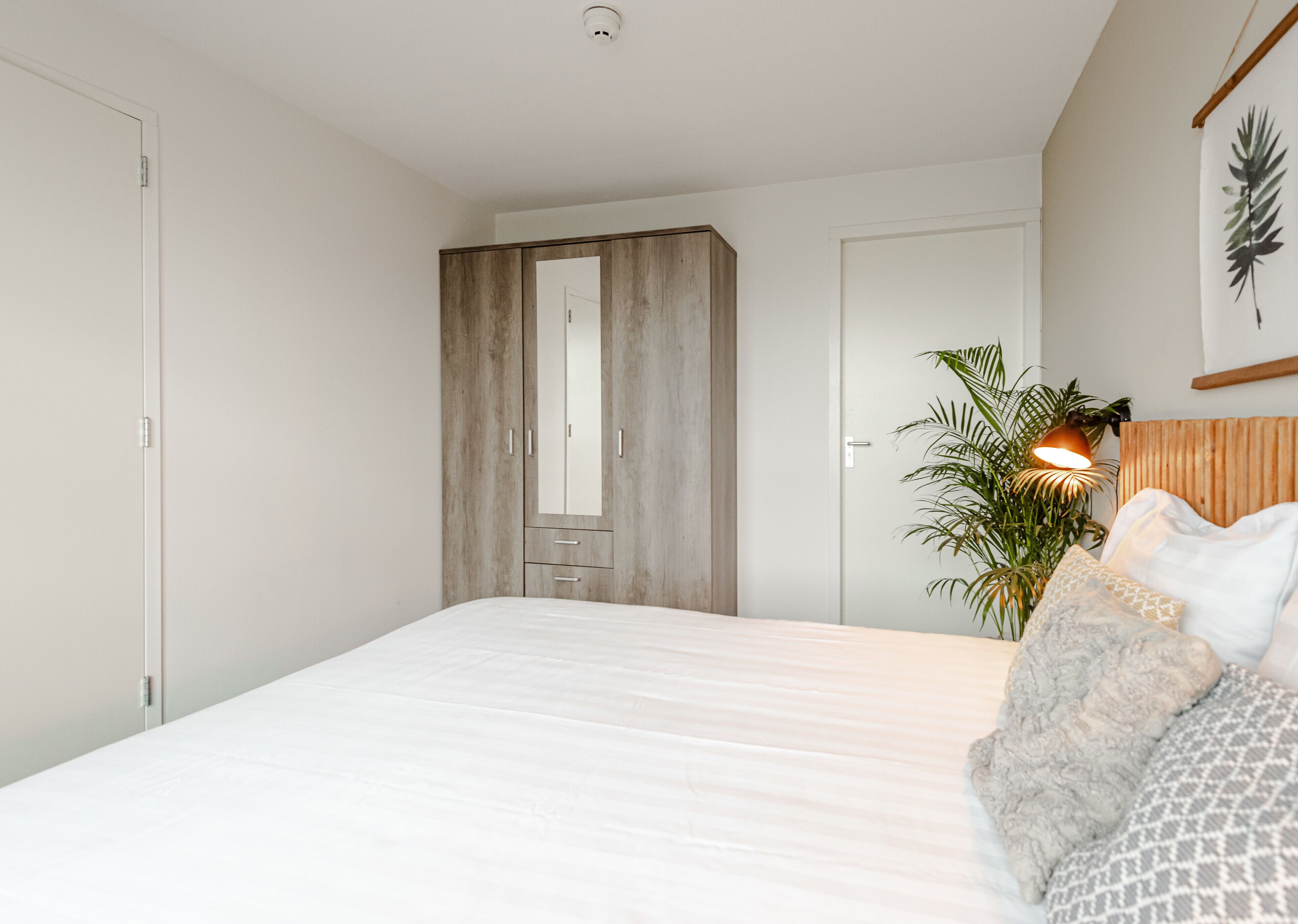 Quarto com uma cama grande, um guarda-roupa, um espelho, uma planta e uma luminária na parede.