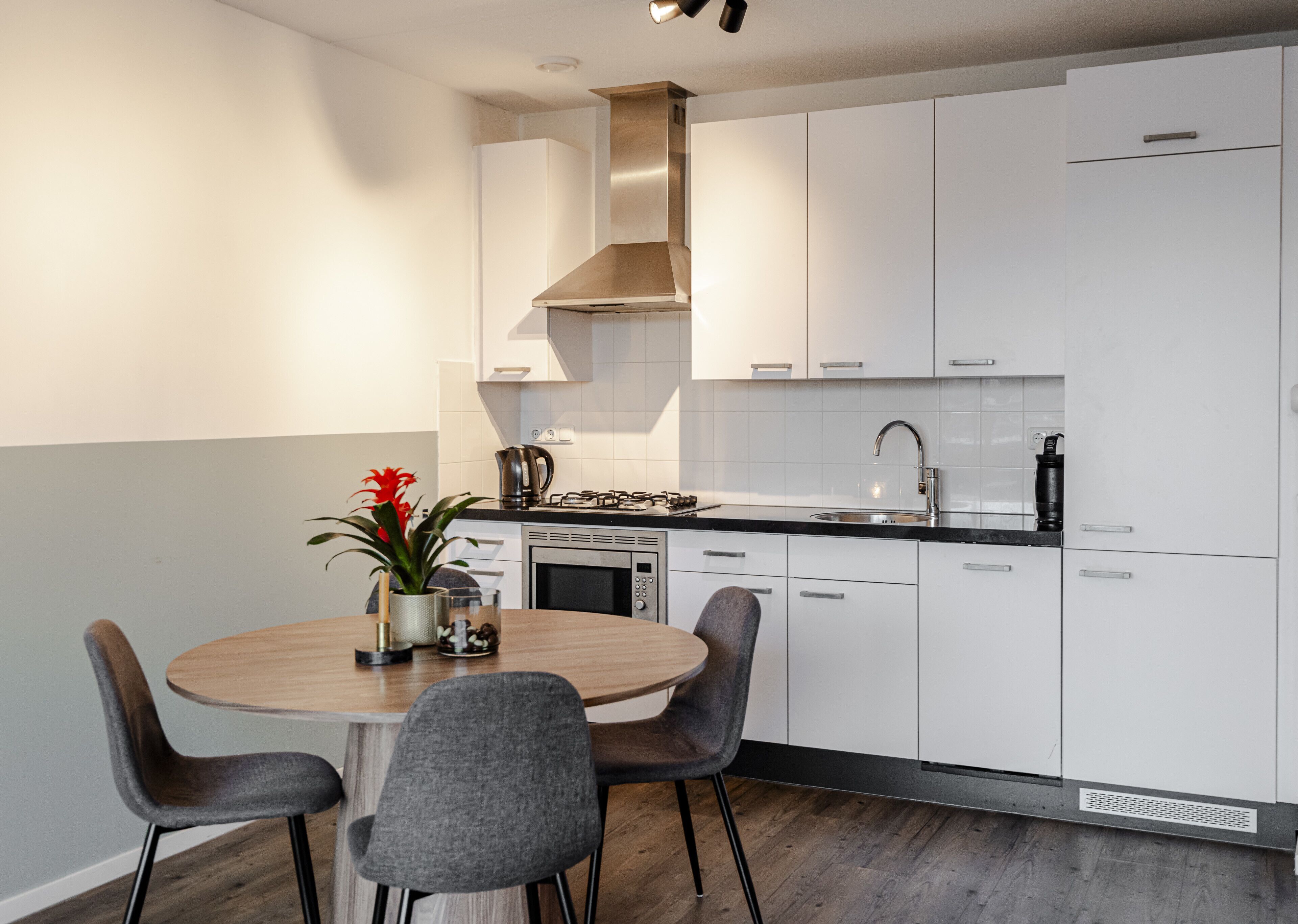 A modern kitchen with white cabinets, a stainless steel range hood, and a black countertop. There is a round wooden table with two chairs and a potted plant.