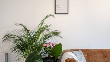 Un salon avec un canapé marron, un tableau encadré au mur et une grande plante verte aux fleurs roses.