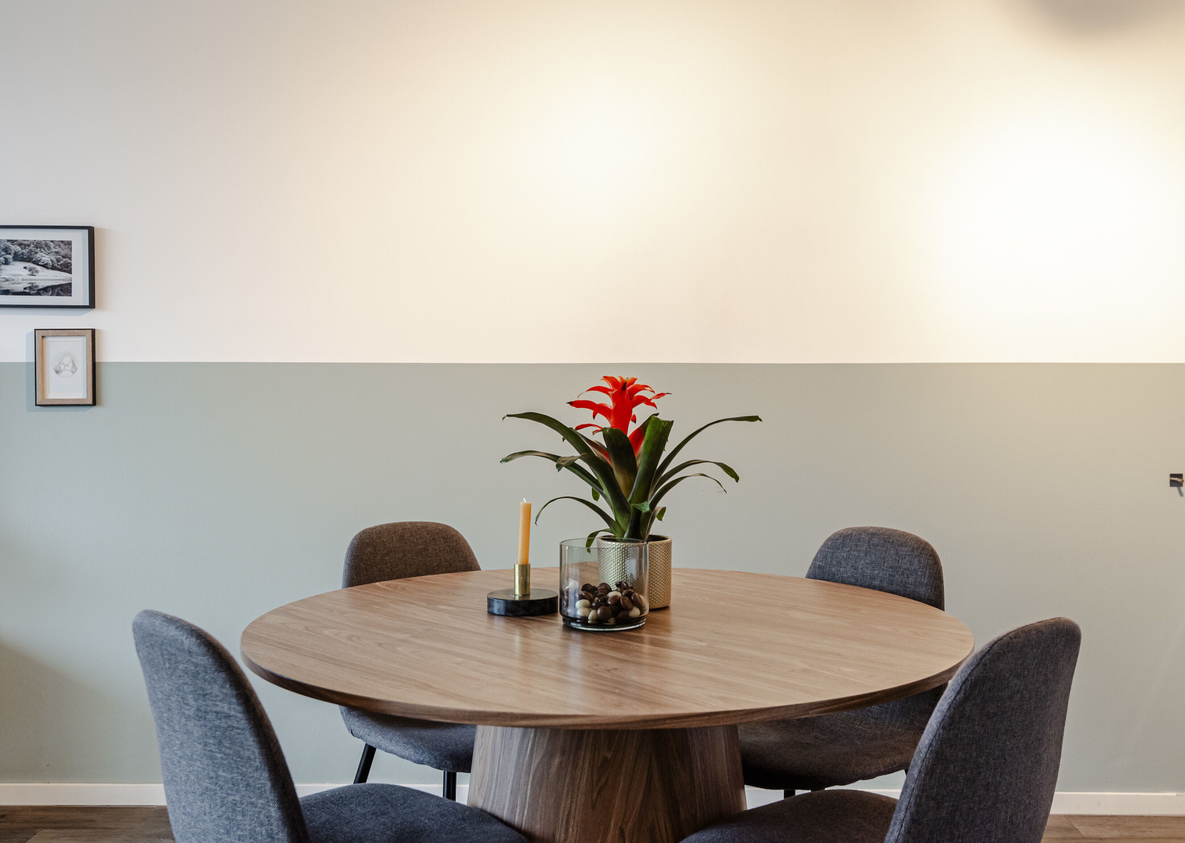 A round wooden dining table with six grey upholstered chairs. A vase with red flowers and a candle are on the table.