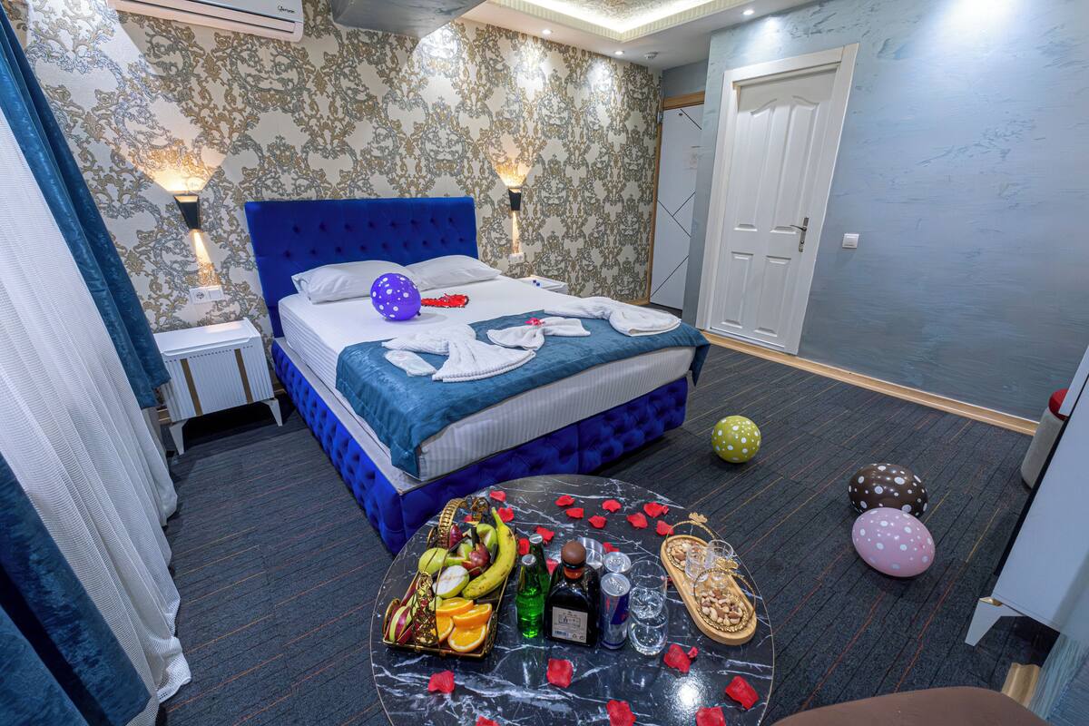 A bedroom with a blue bed, a nightstand, a table with fruits, bottles, and balloons.