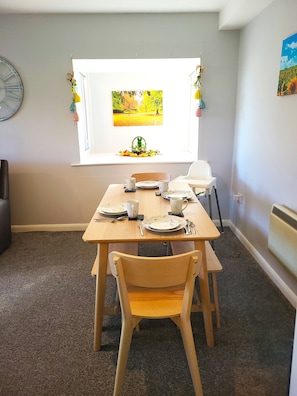 A dining area with a table set for two, a window with a view of a park, and a clock on the wall.