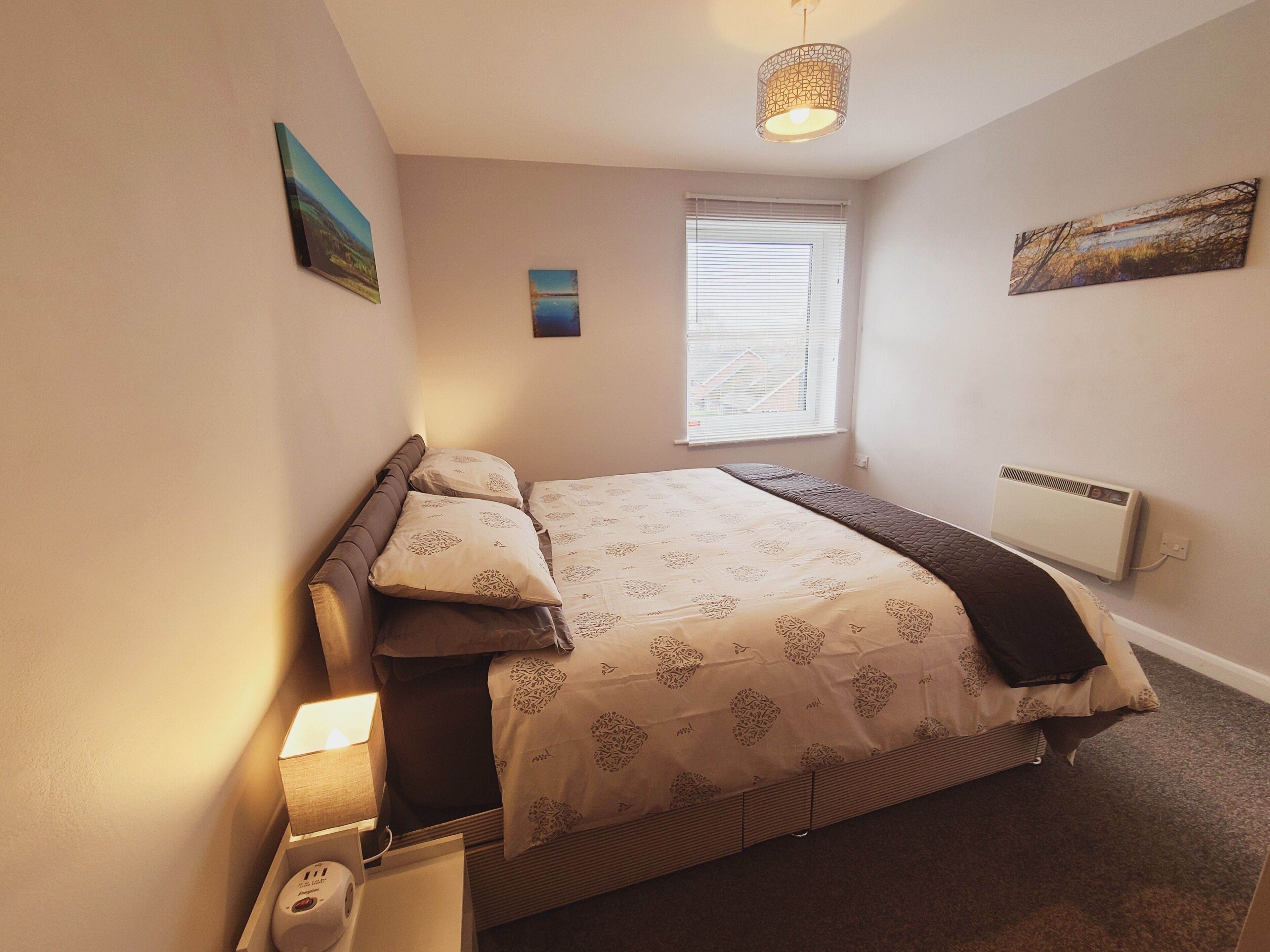 A bedroom with a bed, a bedside table with a lamp, a window with blinds, and a radiator.