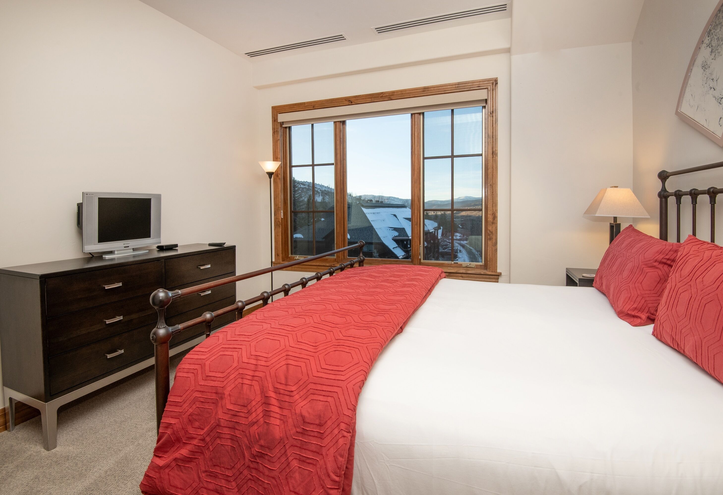 A bedroom with a bed, a dresser, a TV, a window with a view, and a lamp.