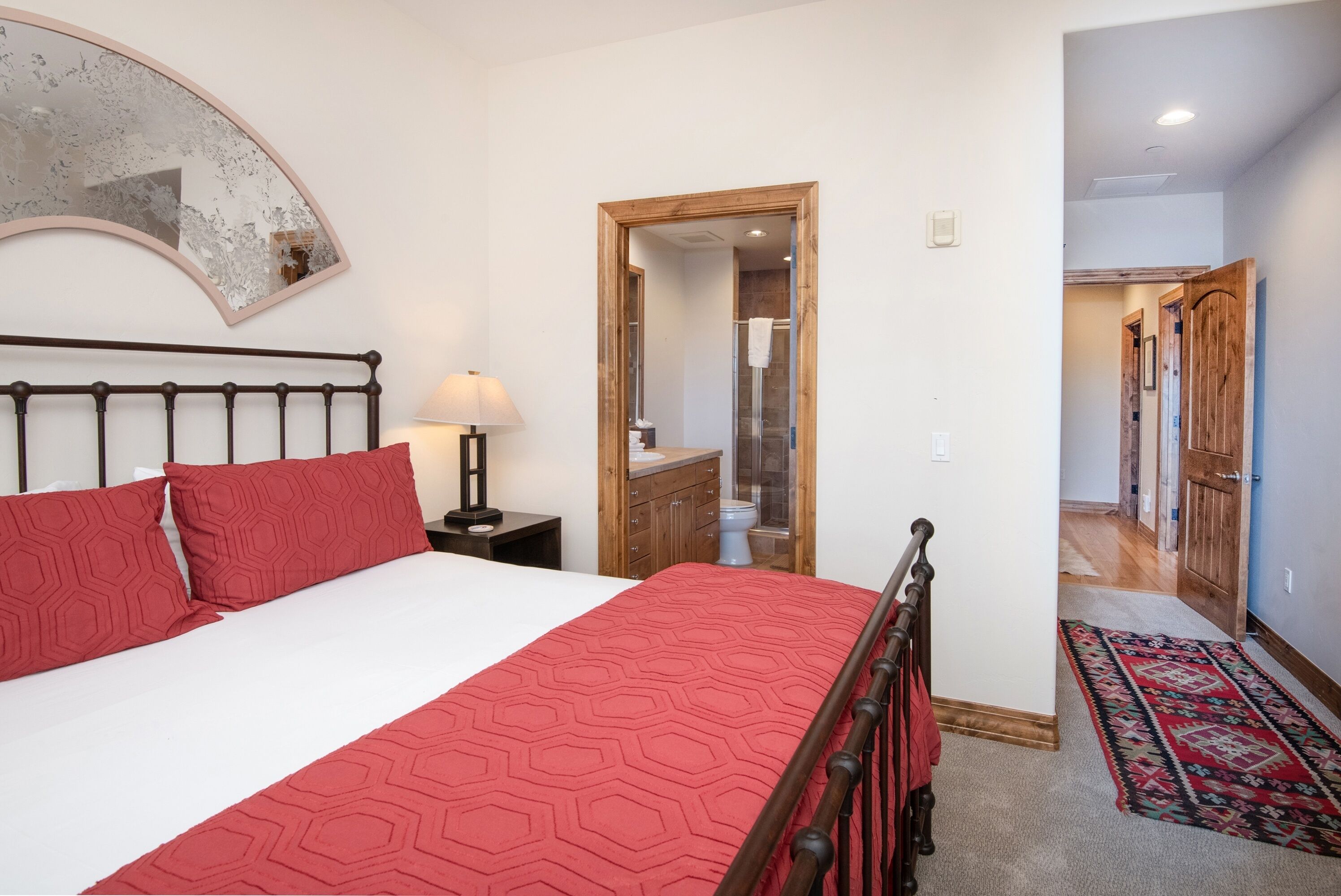 A bedroom with a bed, a bedside table with a lamp, a mirror, and a wooden door leading to a bathroom.
