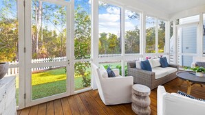 A covered porch with white wicker furniture, a round table, and a view of a yard with a white picket fence and trees. - 107 Sunflower Street 5 Bedroom Home by RedAwning (Santa Rosa Beach)