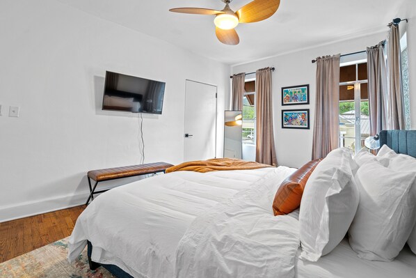 A bedroom with a bed, a ceiling fan, a TV mounted on the wall, and a window with curtains.