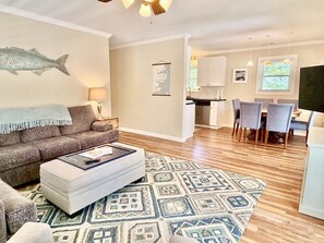 A living room with a sofa, ottoman, patterned rug, and a fish wall art. There is a dining area with chairs and a table, and a kitchen in the background.