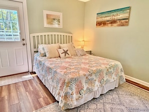 A bedroom with a bed, a door, a window, a wall-mounted picture, and a nightstand with a lamp.