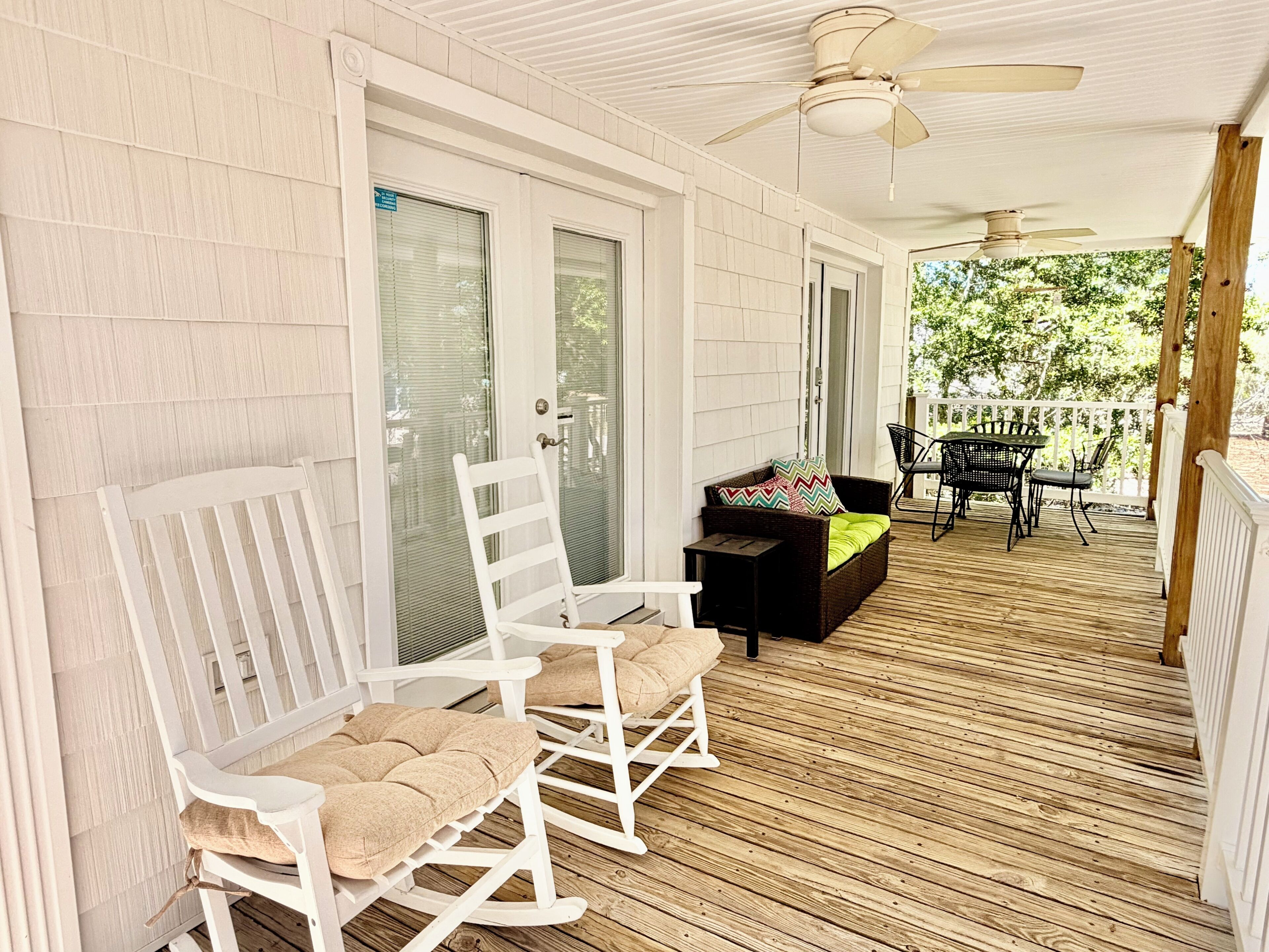 A covered porch with white rocking chairs, a black sofa with a colorful cushion, and a table with chairs.