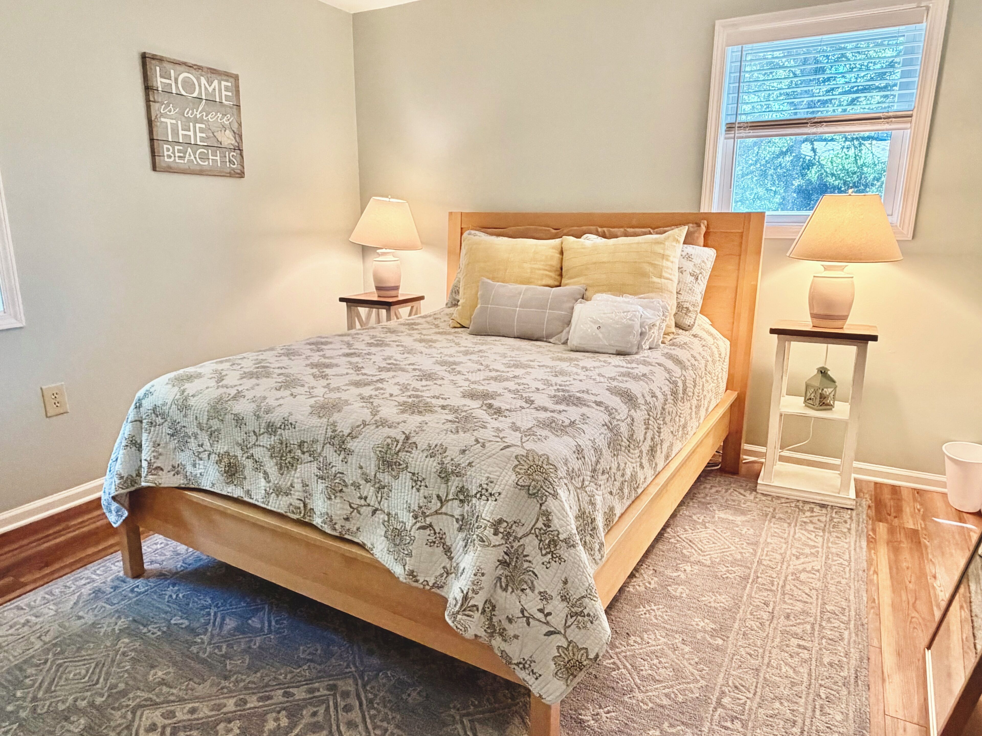 Une chambre à coucher avec un lit, deux tables de chevet avec des lampes, une fenêtre avec des stores, un mur orné d’une inscription « LA MAISON, C’EST LÀ OÙ SE TROUVE LA PLAGE », et un tapis à motifs.