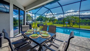 Un espace de restauration extérieur abrité, avec vue sur la piscine, comprenant une table dressée pour quatre personnes, avec des assiettes et un vase de fleurs.