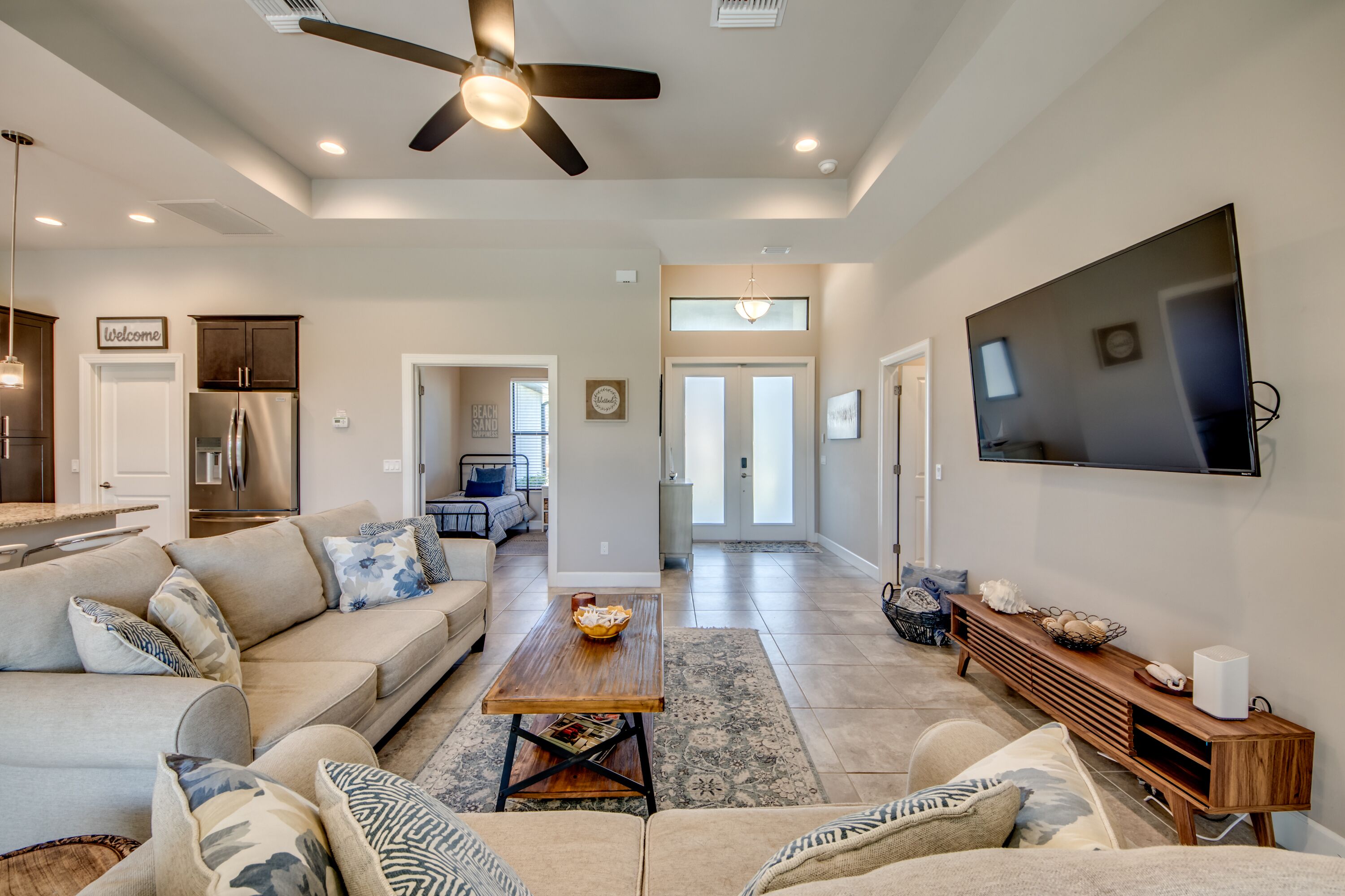 A spacious living room with a large TV, a ceiling fan, and a kitchen in the background.