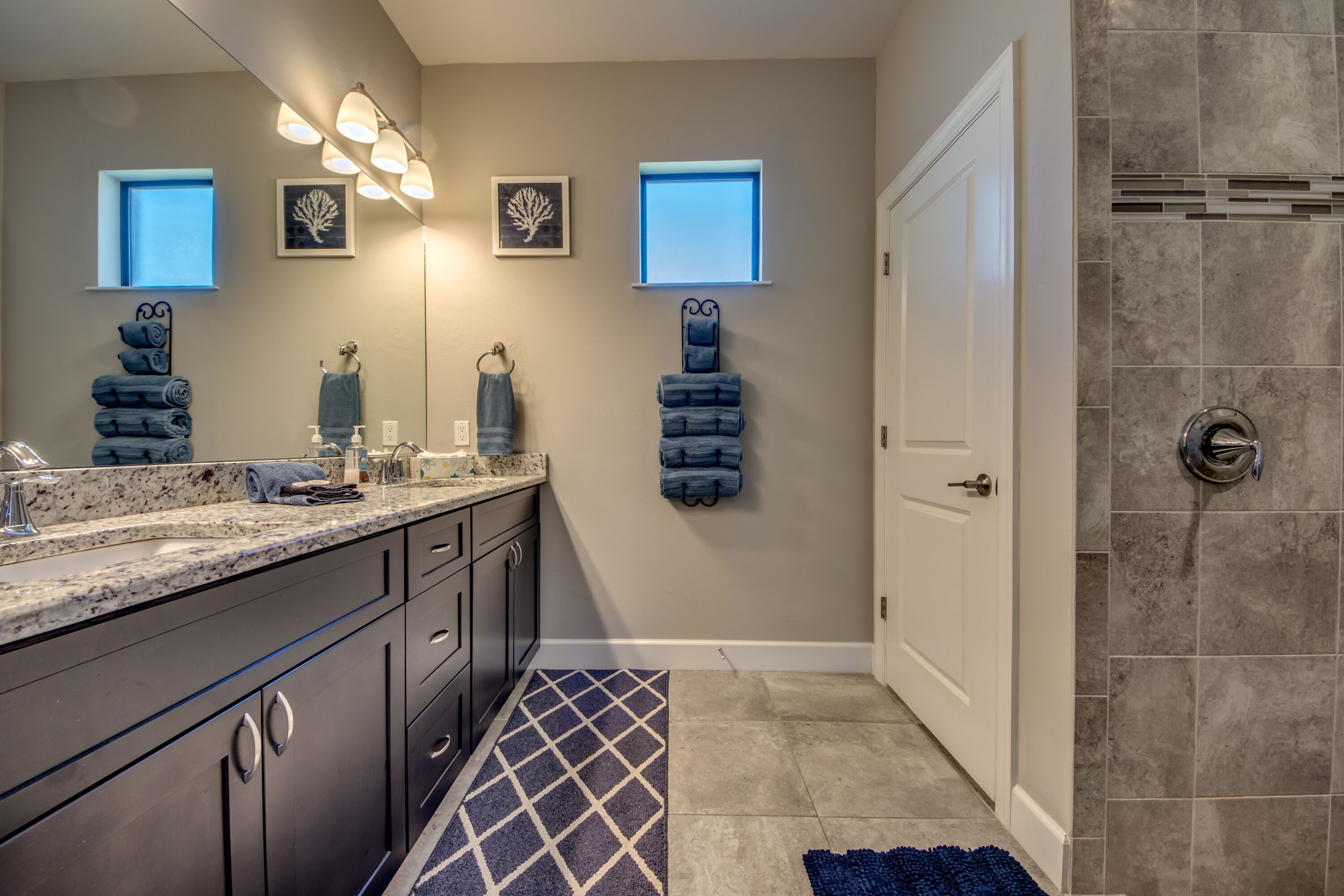 A modern bathroom with a granite countertop, dark cabinets, and a blue patterned rug.