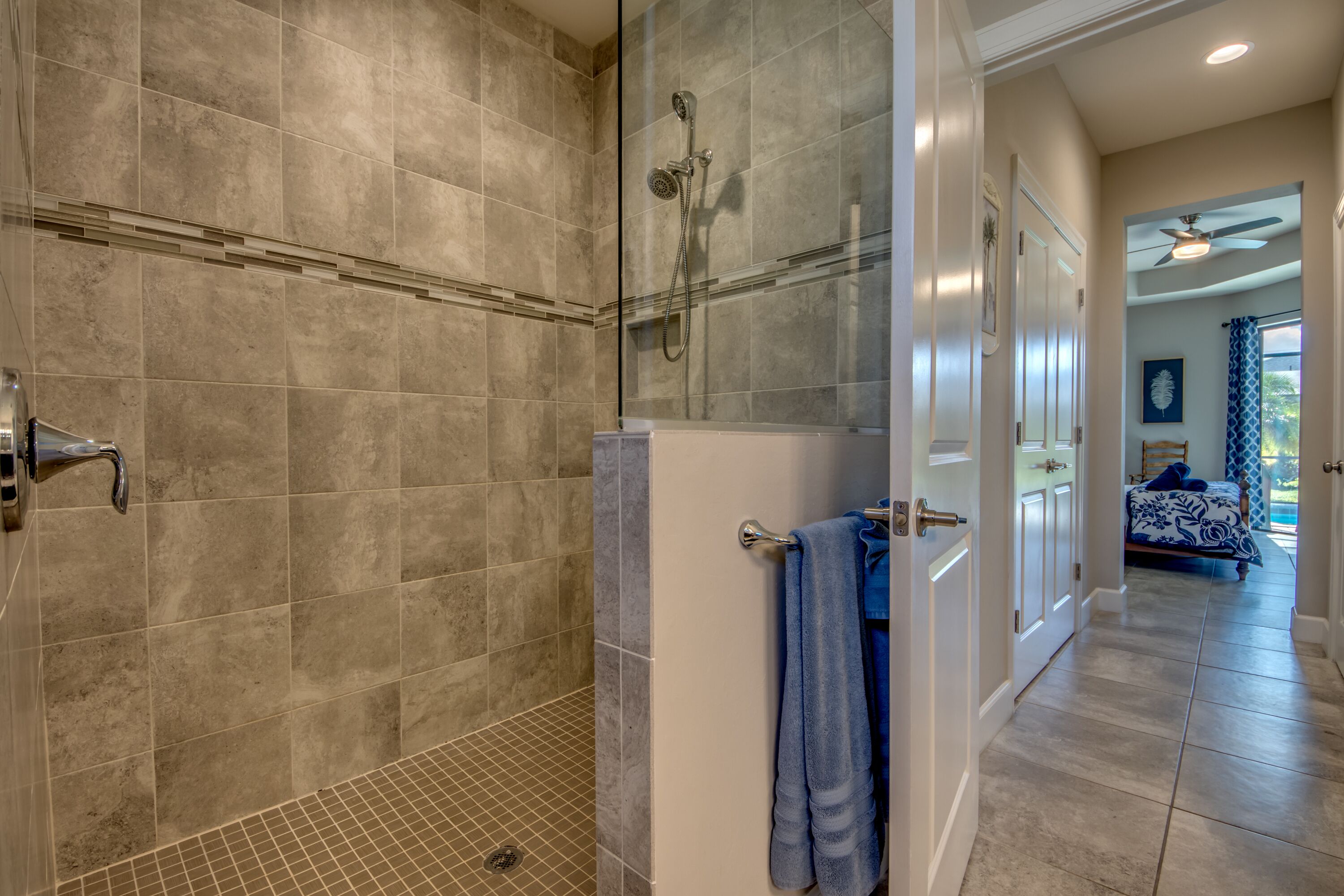 A modern bathroom with a glass-enclosed shower, a visible towel rack, and a door leading to a bedroom with a bed and curtains.