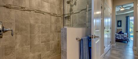 A modern bathroom with a glass-enclosed shower, a visible towel rack, and a door leading to a bedroom with a bed and curtains.