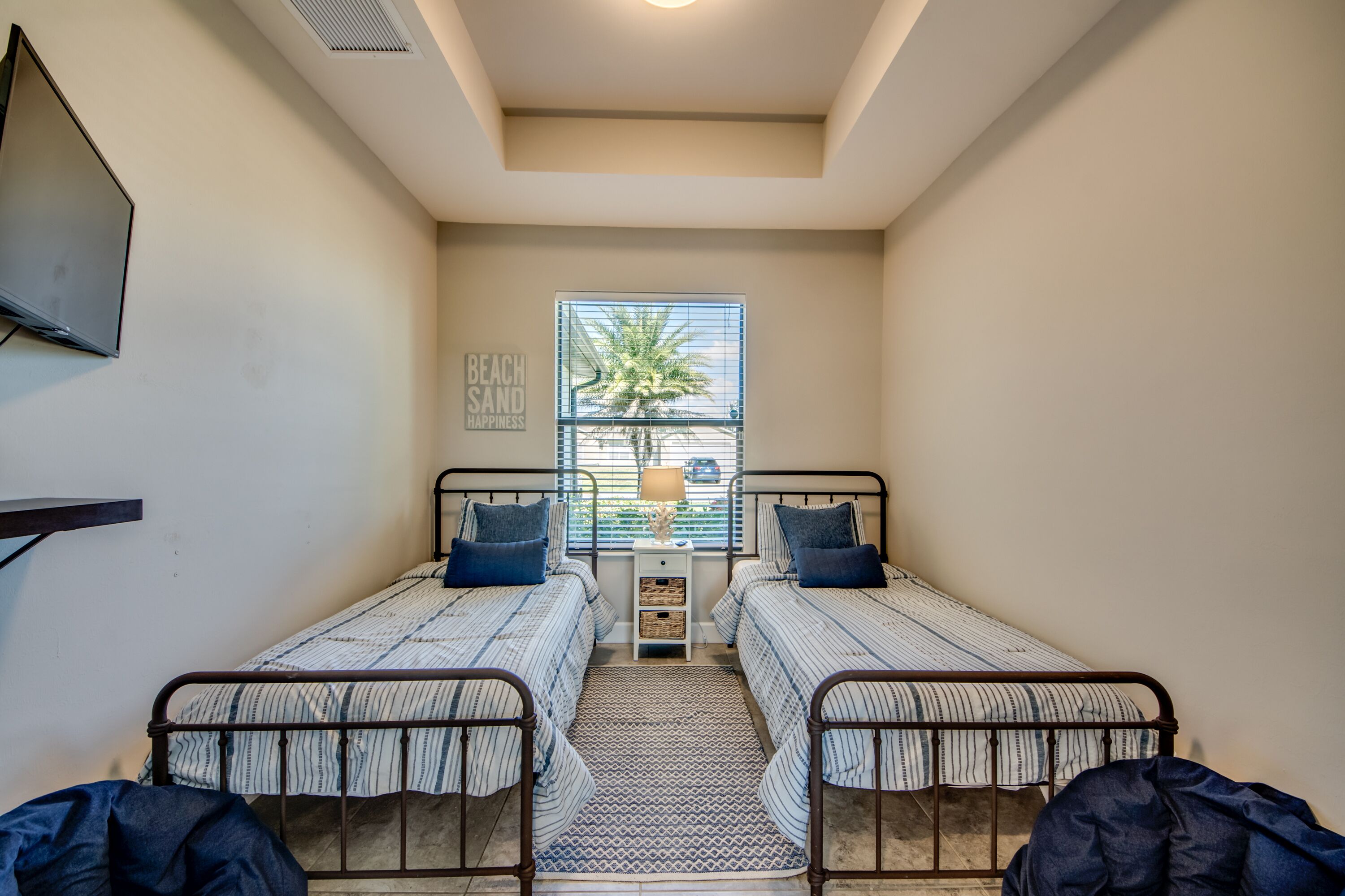 A bedroom with two beds, a window with blinds, a wall-mounted TV, and a small table with a lamp.