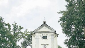 Una església petita de color blanc amb un campanar, envoltada d'arbres i un turó cobert d'herba.
