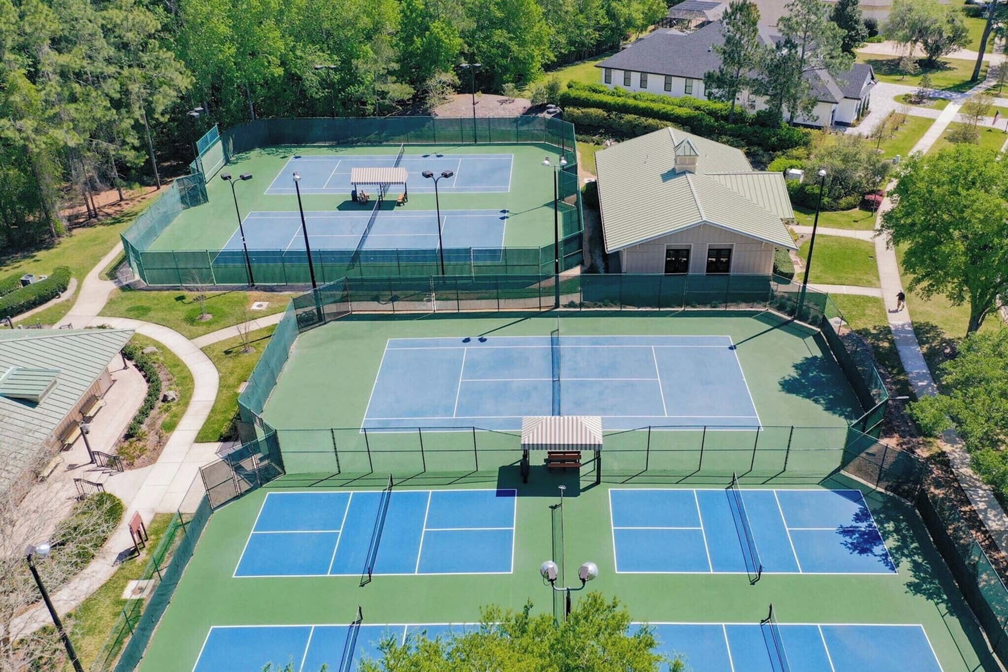 An aerial view of a tennis court complex with multiple courts, surrounded by residential buildings and greenery.